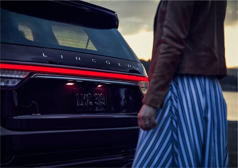 A person is shown near the rear of a 2023 Lincoln Aviator® SUV as the Lincoln Embrace illuminates the rear lights | Purchase Lincoln in Mayfield KY